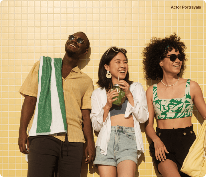 Three friends laughing together against a yellow tiled wall, enjoying a sunny day outdoors.
